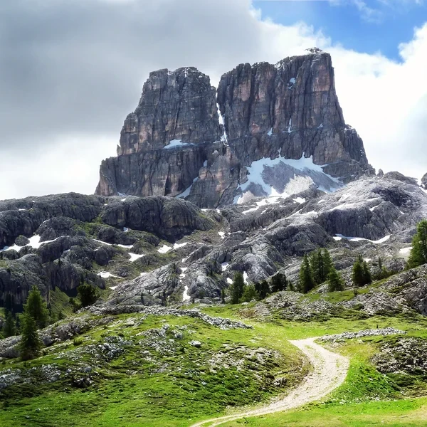 Hiking trail leading toward the towering Averau rock formation with patches of snow on grey limestone slopes and green alpine grass