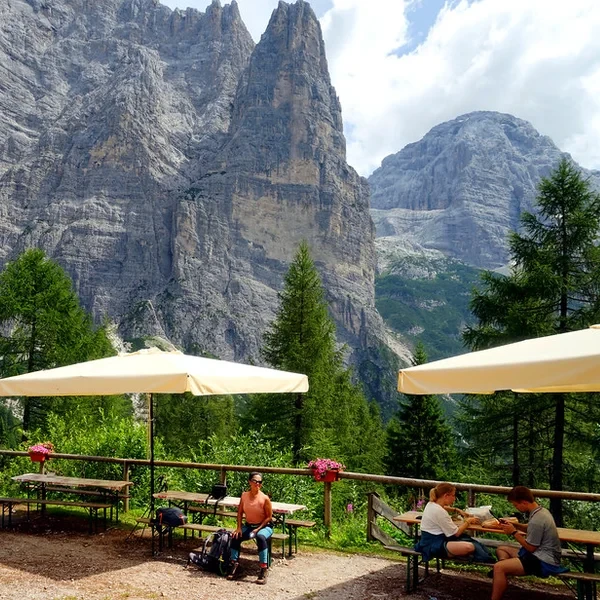 Hikers relaxing on a rifugio terrace with umbrellas and towering Dolomite rock faces behind