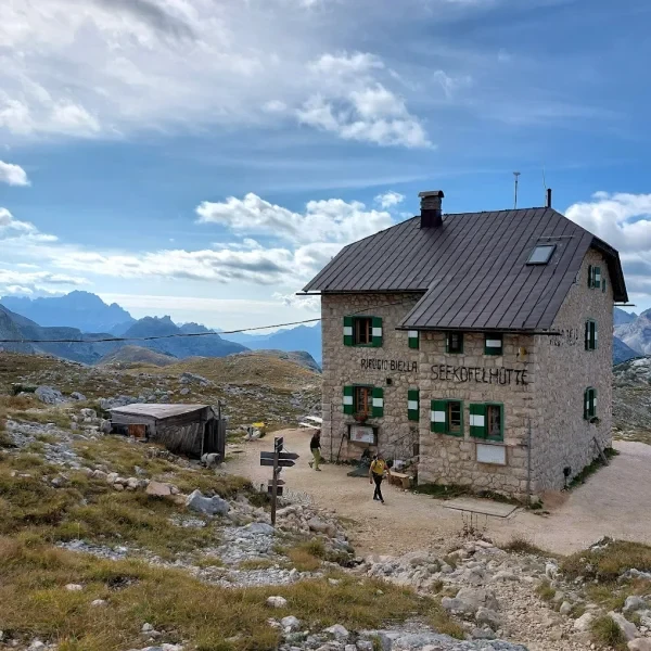 Hikers approaching Rifugio Biella stone hut surrounded by rocky alpine terrain above the Fanes plateau