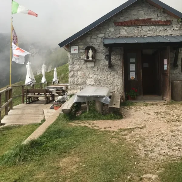 Rifugio Pian de Fontana stone huts on a grassy slope beneath the rugged Schiara group