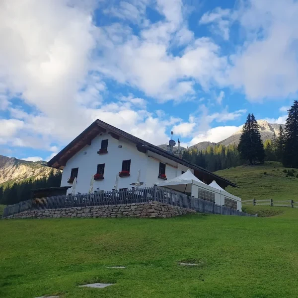 Rifugio Malga Ra Stua alpine hut on a sunny green hillside with wildflowers and mountains behind