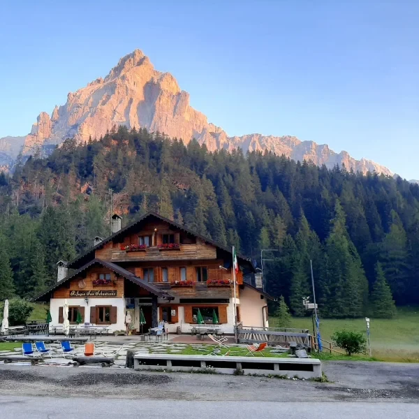 Rifugio San Sebastiano lodge with outdoor seating below a rocky peak and conifer forest