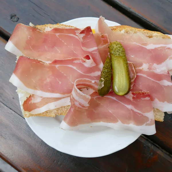 A plate of bread topped with slices of Speck and a gherkin, on a wooden table