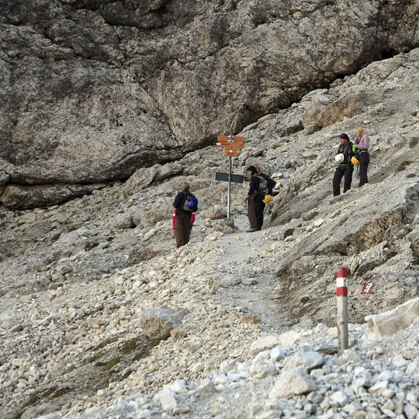 Hikers at a trail junction in front of a signpost, under the rock walls of the Sella group